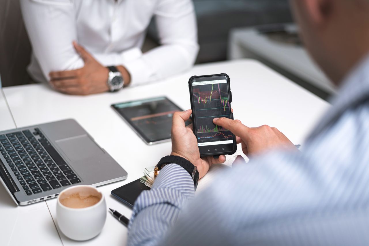 Two businessmen review stock market data on a smartphone and laptop in a contemporary office environment.
